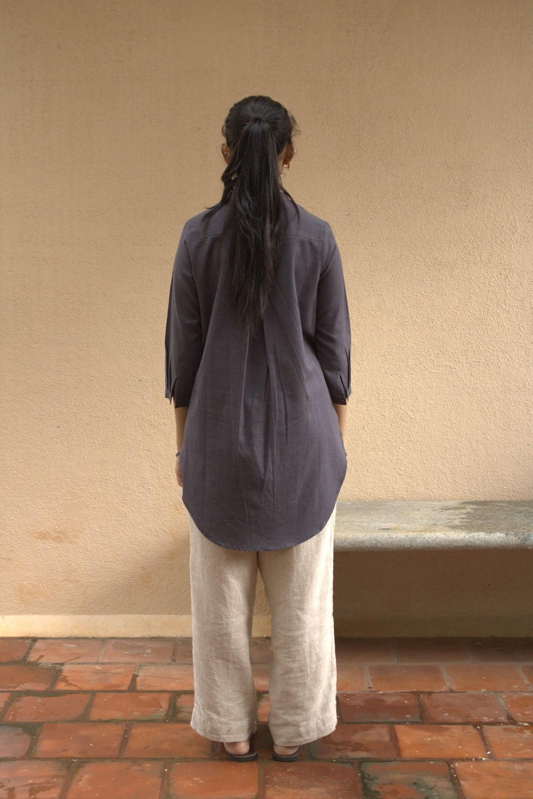 Woman wearing a grey tunic shirt and light pants standing against a beige wall.