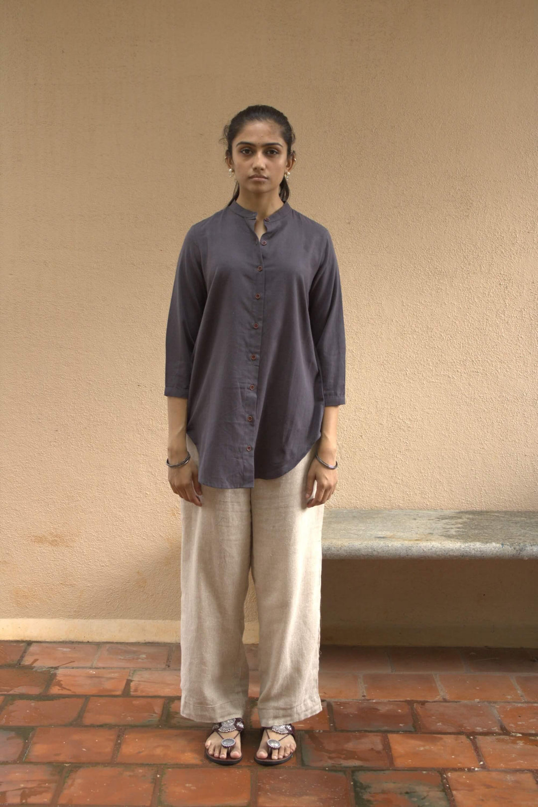 Woman wearing a dark gray tunic shirt and light-colored pants standing against a beige wall.