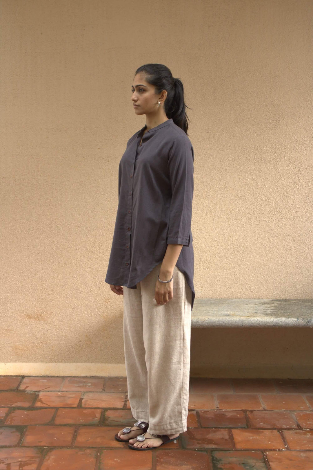 Woman standing against a beige wall with a textured floor, wearing a grey tunic shirt &amp; light colored pants. 