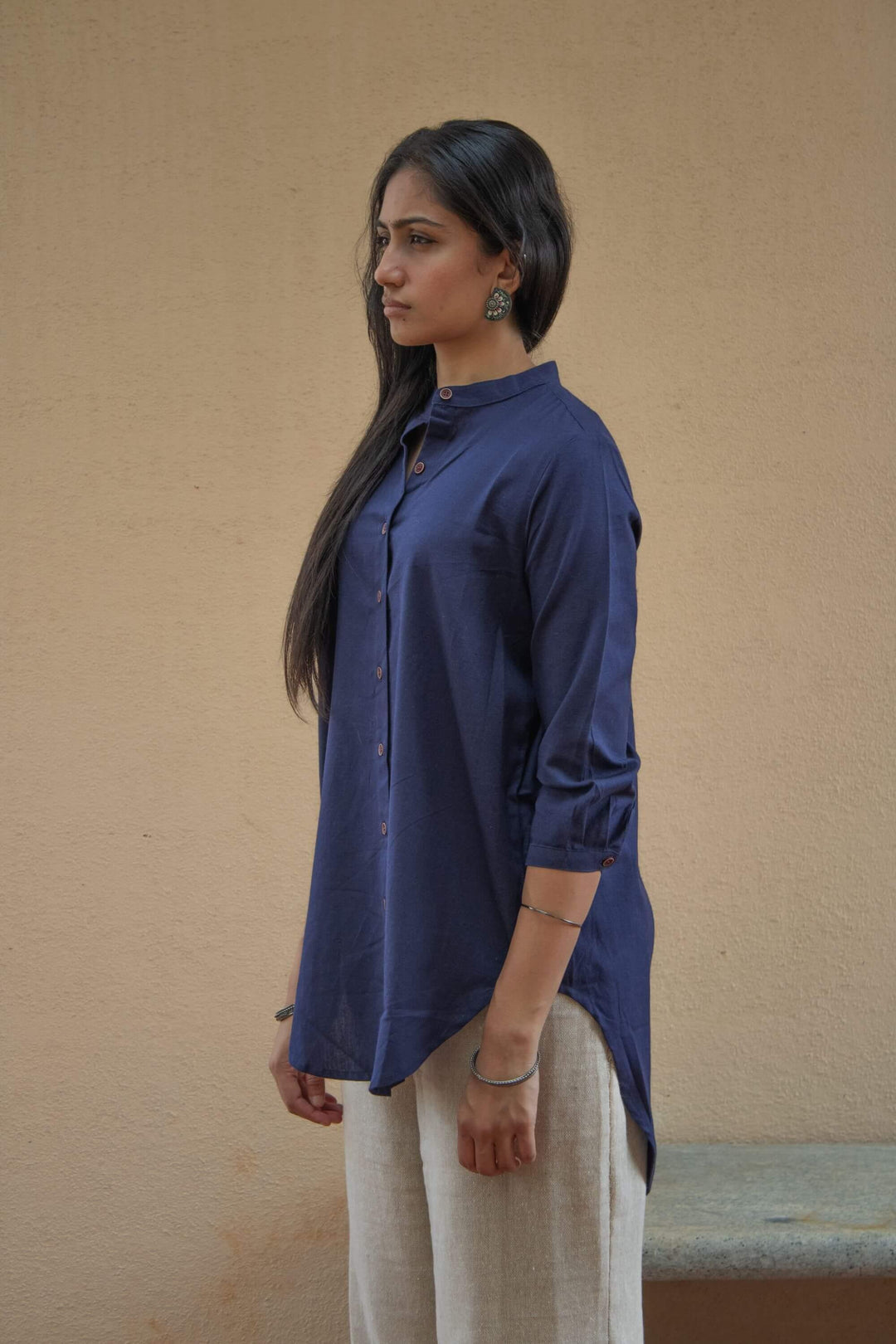 Woman wearing an indigo tunic shirt and beige pants against a beige wall. Side view. 